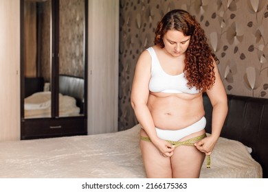 Curly young plus size woman in white underwear stand near bed in bedroom and measure hip circumference with tape. Overweight girls struggle with obesity. Weight loss program, body control and diet.