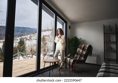 Curly young woman enjoying weekends inside contemporary barn house in the mountains. Happy female tourist looking through panoramic windows in new cottage.