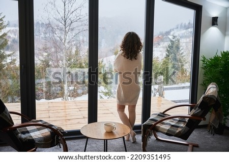 Curly young woman enjoying weekends inside contemporary barn house in the mountains. Back view of female tourist looking through panoramic windows in new cottage.