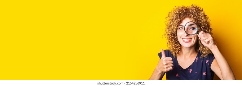 Curly Young Woman Looking At Camera Through Magnifying Glass Showing Thumbs Up Gesture On Yellow Background. Banner