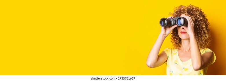 curly young woman looks through binoculars on a yellow background. Banner