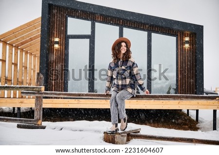 Curly young woman resting on terrace of modern barn house in the mountains. Happy female tourist enjoying winter holidays in new cottage.
