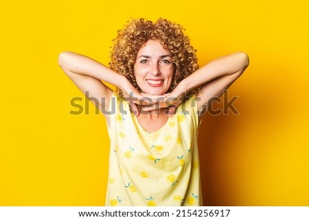 curly young woman smiling holds palms under the chin on yellow background.
