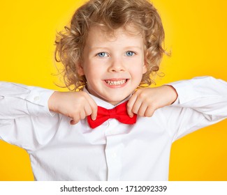 Curly-haired Boy With Blond Hair And Blue Eyes In A Shirt And A Red Bow Tie On A Yellow Background. The Child Is Laughing, Smiling, Having Fun, Happy.