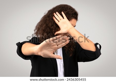 Curly-haired businesswoman in blazer making decisive stop gesture with her hand and covering face, indicating boundary or refusal, on grey background