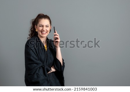 curly-haired emotional young woman in a black cloak using mobile phone, isolated on gray background