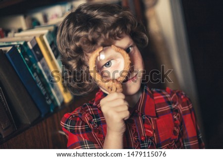 A curly-haired girl in a checkered red dress stands in front of a bookshelf and holds a magnifying glass in front of her face.