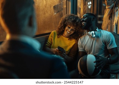 Curly-haired girl looking at the smartphone in her hands and laughing with a young black man holding a basketball on a crowded bus at night. - Powered by Shutterstock