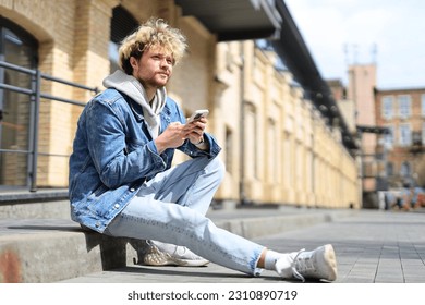 Curly-haired Man sits on the curb with a phone in his hands. - Powered by Shutterstock