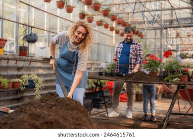 Curly-haired mother playfully shovels soil in the greenhouse while her husband and daughter plant flowers, a moment of family fun and learning. - Powered by Shutterstock