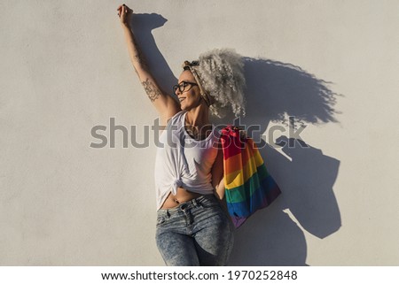 A curly-haired Spanish cute woman with a pride flag tote bag and tattoos on a white wall background
