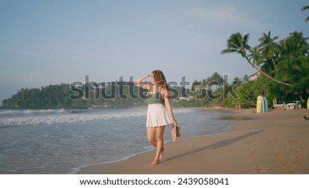 Curly-haired woman strolls on tropical beach. Solo traveler enjoys serene sea coast, morning walk. Leisure vacation, tranquil oceanfront promenade. Female explores sandy shore, summer vibe.