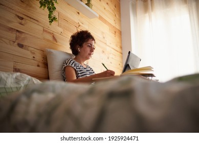 A Curly-haired Woman Takes Notes In Her Notebook While Telecommuting From Her Bedroom. The Room Has A Nice Wooden Wall And It Is Daytime. New Technologies Allow You To Work Comfortably.