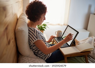 A Curly-haired Woman Takes Notes In Her Notebook While Telecommuting From Her Bedroom. The Room Has A Nice Wooden Wall And It Is Daytime. New Technologies Allow You To Work Comfortably.