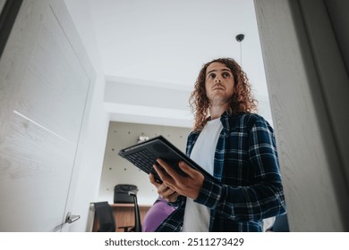 Curly-haired young man stands in an office environment, holding a digital tablet thoughtfully, dressed in a casual blue plaid shirt. - Powered by Shutterstock