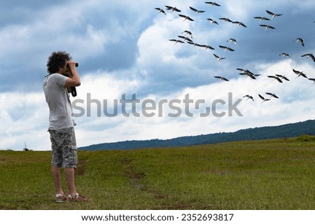 Curly-haired young man watches migrating storks with binoculars.  Nature watcher person. Birdwatching, zoology, ecology. Research in nature, observation of animals. Ornithology. Horizontal photo. 