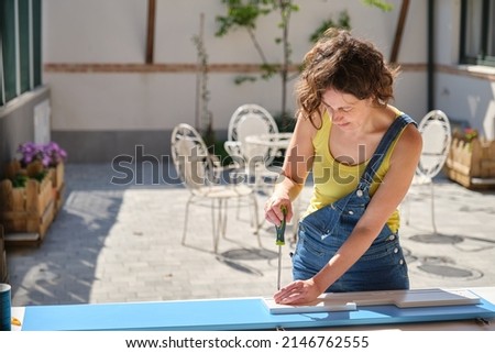 curly-haired young woman in blue and yellow clothes in the sunlight in the garden assembling a blue and white piece of furniture with a screwdriver