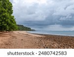 A curved beach under glowering overcast skies with tropical rainforest growing up to the edge of the beach which is strewn with stones exposed by the tide at Mission Beach in Queensland, Australia.