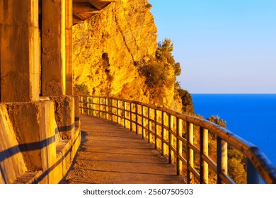 Curved cliffside pathway with metal railings, illuminated by golden sunset light, contrasts against a vibrant orange rock face and a deep blue sea under a clear sky. Walking path along a cliff - Powered by Shutterstock