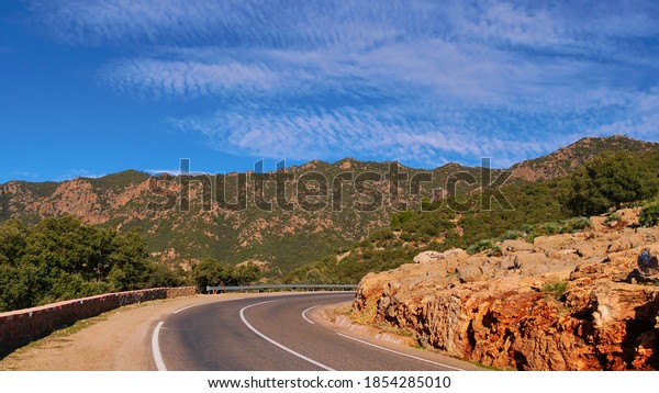 Curved paved road with crash barriers leading\
through the foothills of Altas Mountains south of Beni Mellal,\
Morocco with hills covered by forest on a sunny winter day with\
blue sky and fleecy\
clouds.