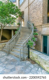 Curved Staircase At The Exterior Of An Old Building With Brick Wall. The Outside Stairway Has Metal Railings Decorated With Hanging Plants With Small Colorful Flowers.