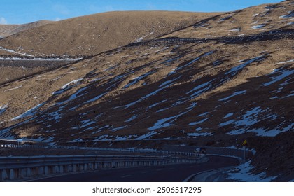 Curving road winds through a barren, snow-dusted hillside under a vast blue sky, creating a contrast between the rugged terrain and open space. - Powered by Shutterstock