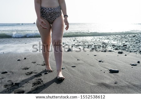 Curvy adolescent walking in the beach in the sunset in the wet black sand and waves on the sea. Caucasian woman in bikini at the coast, with barefoot and pleasure.