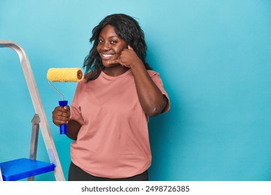 Curvy woman with paint roller and ladder showing a mobile phone call gesture with fingers. - Powered by Shutterstock