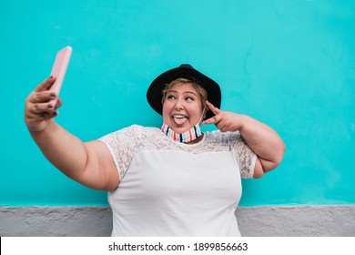 Curvy woman wearing protective face mask under chin and taking selfie with mobile phone outdoor - Coronavirus lifestyle and technology trends - Focus on the face - Powered by Shutterstock