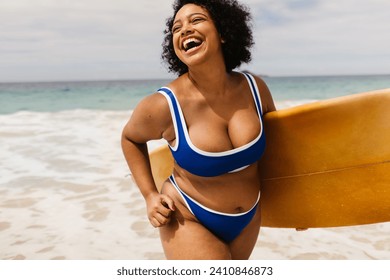 Curvy young woman enjoys beach activities, celebrating summer on a solo vacation. Female surfer carrying a board and wearing a bikini, capturing the essence of a fun and authentic summer holiday. - Powered by Shutterstock