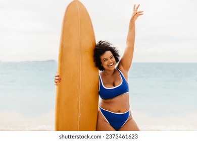 Curvy young woman holds her surfboard and raises her arm in celebration after an exciting surfing adventure on a sunny beach. Happy plus size woman exploring beach activities in a bikini. - Powered by Shutterstock