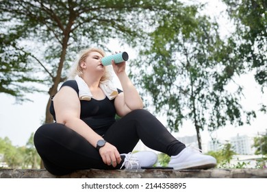 Curvy Young Woman Sitting On Stone Curb In Park And Drinking Electrolyte Water After Jogging In Park