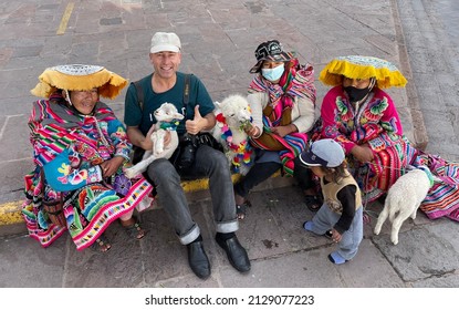 Cusco, Peru, 21 November 2021: Happy Male Tourist Sitting With Indigenous Peruvian People In Traditional Clothes And Holding Little Alpaca