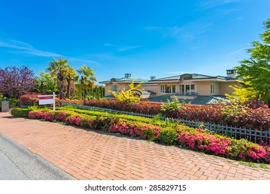 Custom Built Luxury House With Nicely Trimmed And Landscaped Front Yard Lawn And Driveway To Garage In A Residential Neighborhood. Home For Sale Sign From Real Estate Agency.