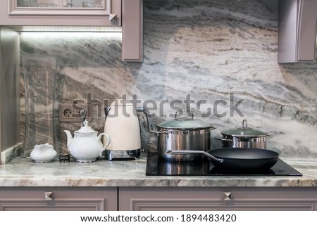 custom designed kitchen, with marble looking quartz countertop and backsplash. cream electric kettle with porcelain tea accessories on granite countertop next to ceramic hob with steel pots and pan