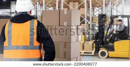 Custom warehouse. Man customs officer and boxes. Guy in yellow vest with her back to camera. Tiered racks and forklift blurred. Customs warehouse with cardboard boxes. Career storage worker.