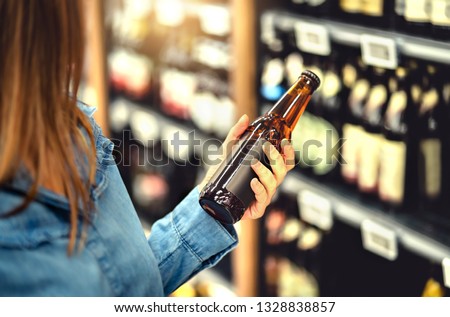 Customer buying beer in liquor store. Lager, craft or wheat beer. IPA or pale ale. Woman at alcohol shelf. Drink section and aisle in supermarket. Lady holding bottle in hand. Drink business concept.
