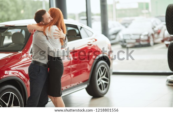 Customer, car sale, consumerism and people\
concept - laughing cheerful couple embracing on the new red car\
background at car\
showroom
