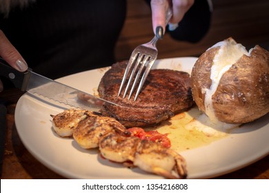 Customer Is Cutting The Steak , With Baked Potato And Grilled Shrimp In White Plate.