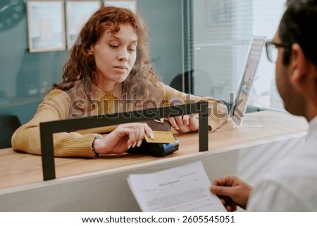 Customer engaging with bank representative while processing financial transaction at service counter with paperwork and credit card reader visible on desk