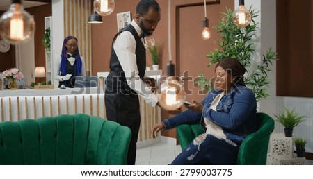 Customer makes nfc payment at pos to pay for coffee cup while waiting on accommodation vacancy in five star hotel, African American traveller talking to bellboy about luxurious service.