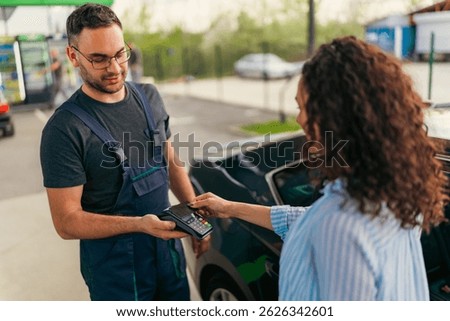 Customer making a contactless payment using her smartphone at a gas station, while a mechanic holds the pos terminal, providing convenient and modern transaction