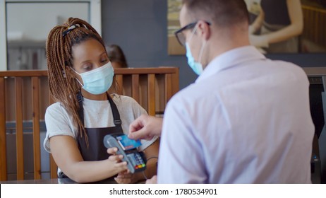 Customer Paying With Credit Card For Buying Food On Counter In Modern Cafe Wearing Safety Mask. African Waitress Holding Card Reader For Client To Pay For Order In Coffee Shop.