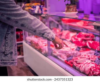  customer is pointing with their finger at the products displayed on the counter of a butcher shop. Their hand and arm are in closeup, highlighting the variety of fresh and selected meat  - Powered by Shutterstock