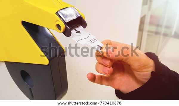 Customer pulls with hand a numbered ticket\
out of yellow number dispenser machine, to wait in service line and\
to be served when his number is\
displayed