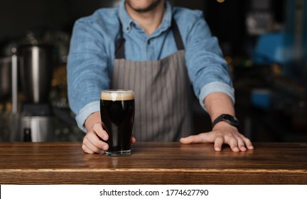 Customer service during party. Barman in denim shirt and apron stands behind wooden counter and gives glass of dark beer, cropped - Powered by Shutterstock