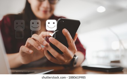 Customer service evaluation concept. smiling Asian female Is using a smartphone And she is pressing face emoticon smiling in satisfaction on virtual touch screen.
