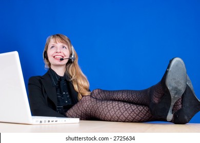 A Customer Service Representative Smiling During A Telephone Conversation With Her Feet On The Desk, Shot Against A Dark Blue Background