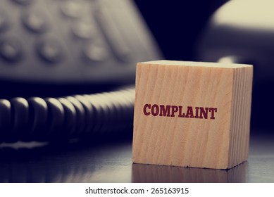 Customer Services Complaint Concept With A Small Wooden Cube Bearing The Word - Complaint - Standing On A Desk Alongside A Land Line Telephone Instrument In A Close Up View.