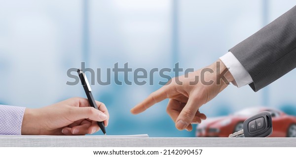 Customer signing a car purchase agreement, the\
salesman is pointing at the\
contract